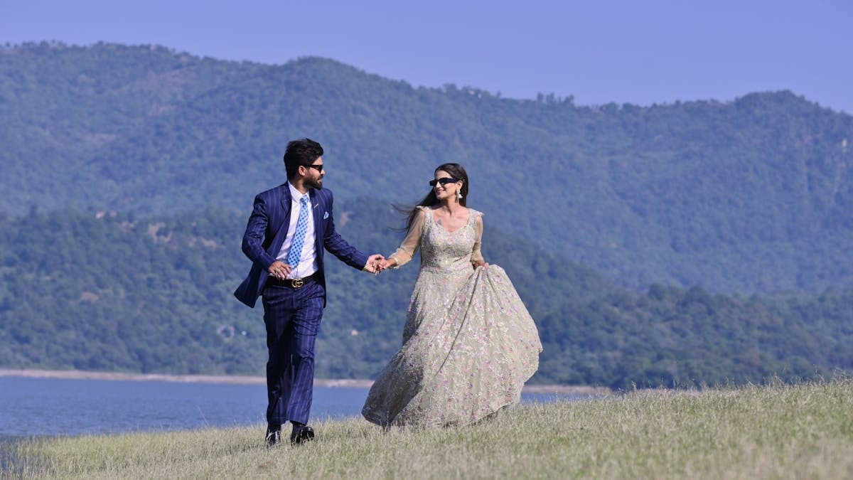 A couple in elegant wedding attire walking hand in hand by a picturesque riverbank in Una, India.